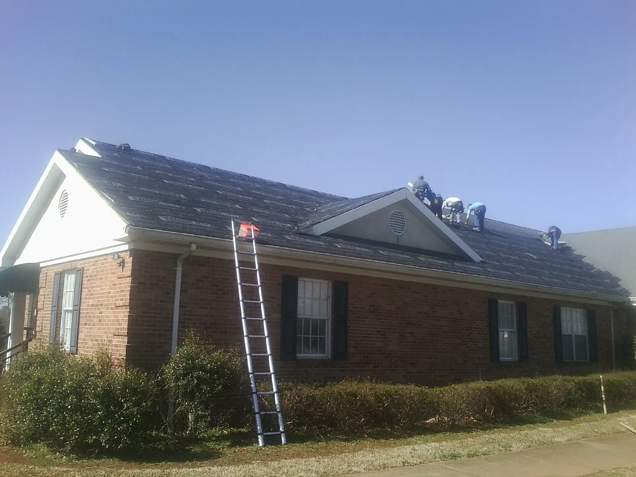 Crew tearing shingles off a brick ranch roof with a ladder set — Emergency Roof Repair work by our crew in Ace