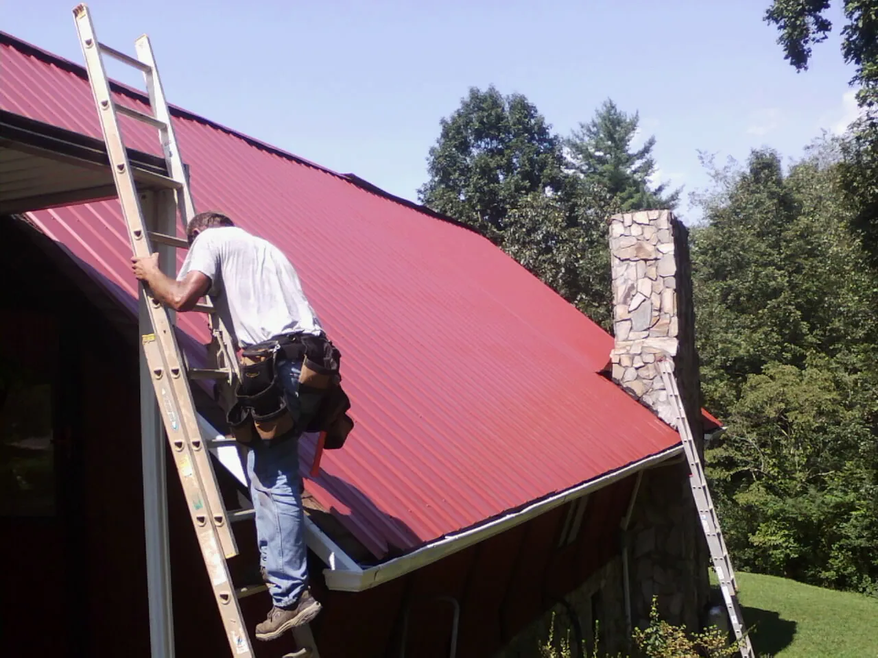 Roofer on a ladder beside a red metal roof and stone chimney — Soffit & Fascia Repair by Roofing Brothers in Ace